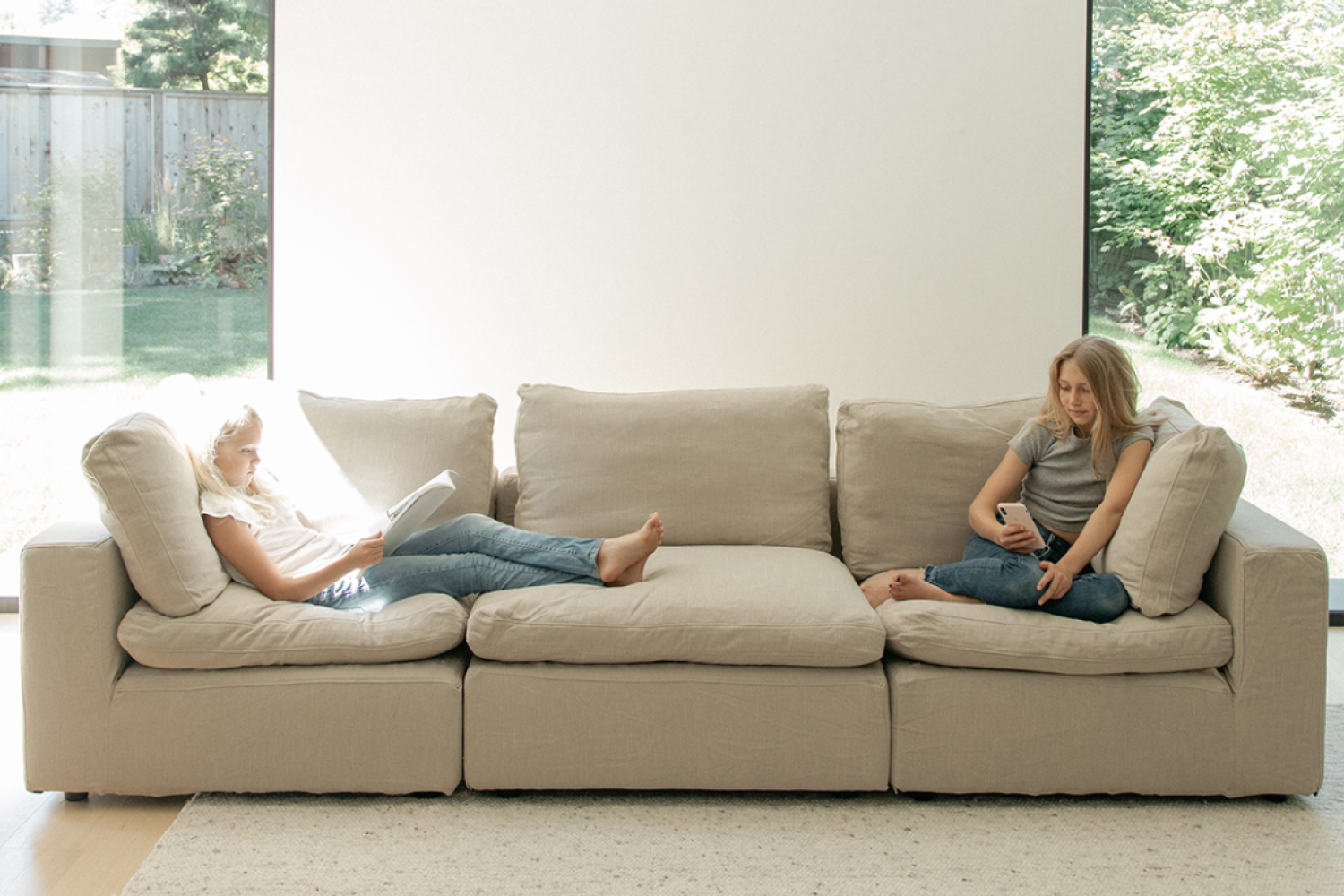 Two girls are comfortable lounging on an EM Sky Sofa in a well-lit living room with a clear view of a green backyard through a floor-to-ceiling window.