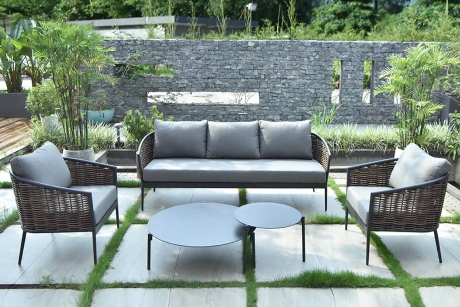 Malcom Grey 5-Piece Wicker Patio Sofa Set arranged on a garden patio, including a sofa, two chairs with cushions, and two round coffee tables, against a backdrop of lush greenery and a stone wall.