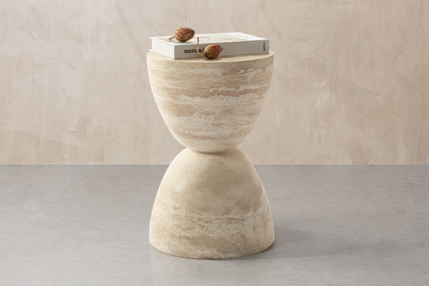 A unique Sera Hourglass White Travertine Side Table table with a textured design resembling an hourglass, topped with a small book and two acorns, against a simple and elegant beige backdrop.