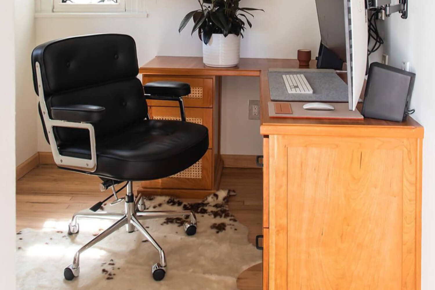 A modern Executive Chair with black upholstery and silver metal accents, positioned at a wooden desk with a computer setup, tablet, and a potted plant within a bright home office space.