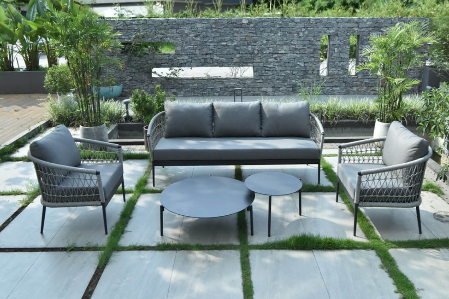 Inviting outdoor patio space with Duxton Grey 5-Piece Rope Weave Patio Sofa Set. The area is surrounded by lush greenery, potted plants, and a stylish stone wall. 