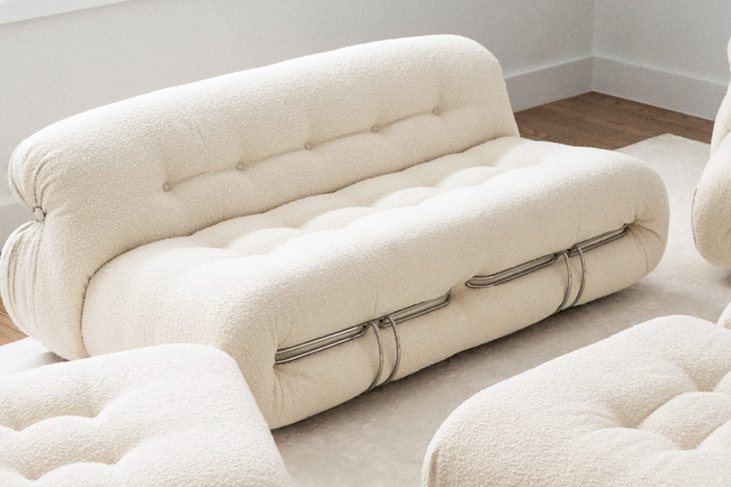 The Soriana Two-Seater Sofa upholstered in a soft, creamy fabric with deep tufting and unique chrome clasps that embrace its plush cushions, creating a luxurious and inviting seating experience.