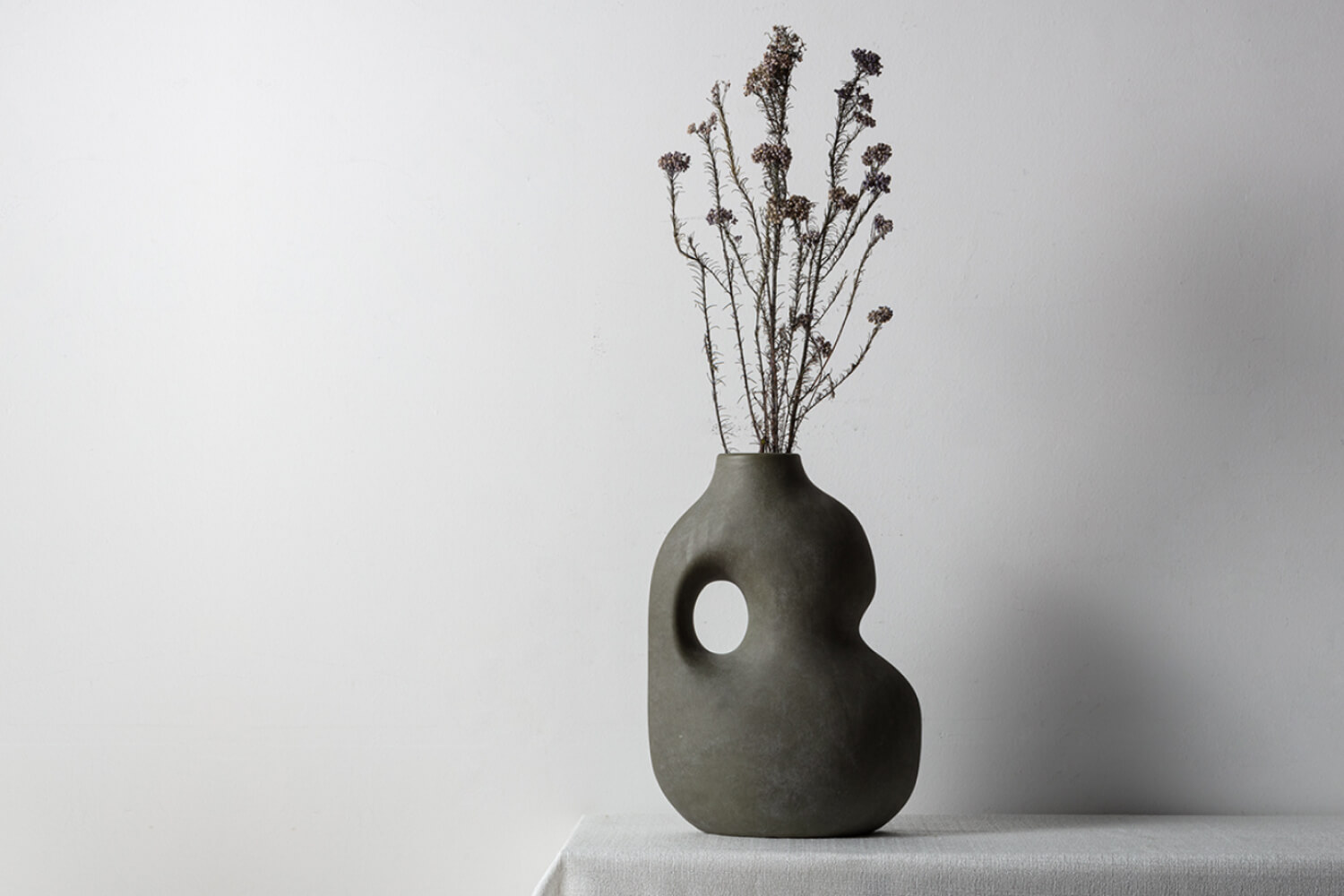 Willow Decorative Sculptural Faded Earth Grey Rustic Wabi Sabi Ceramic Vase set against a neutral wall on a draped tablecloth.