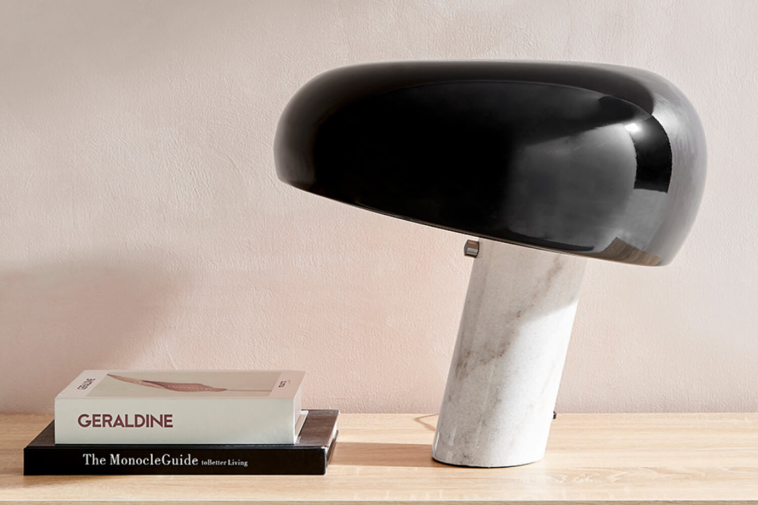 A Snoopy Table Lamp with a distinctive black hood-like shade and white marble base, styled next to a book on a wooden surface against a soft peach-colored wall.