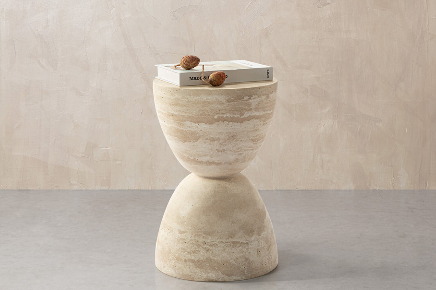 The Sera Hourglass White Travertine Side Table showcases a natural stone finish with layered textures, topped with a book and decorative items.