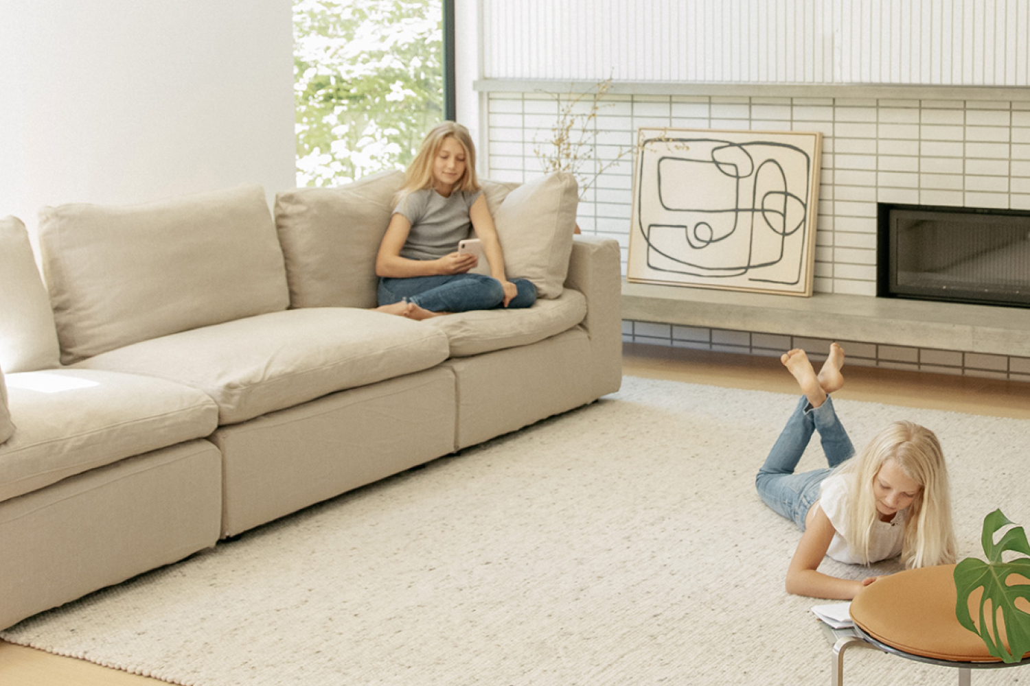 A comfortable and modern living room scene with two girls. One is sitting on a large beige EM Sky Sofa, focused on her smartphone, and the other is lying on the floor, reading a book.