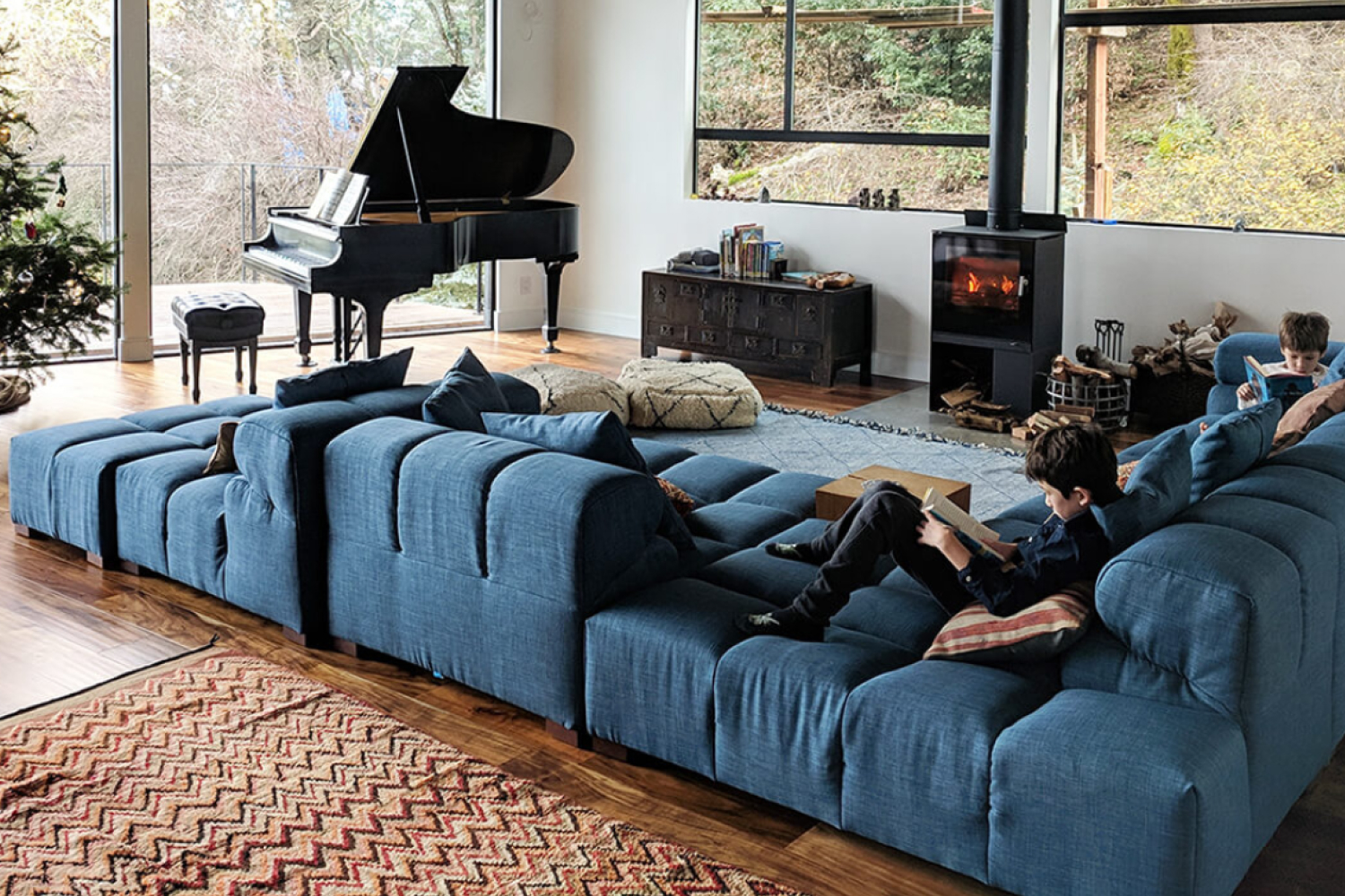 A cozy living room scene with two boys relaxing on a Tufted sofa, each engrossed in reading a book. A grand piano sits near large windows offering a view of trees, and a warm fireplace adds to the room's ambiance.
