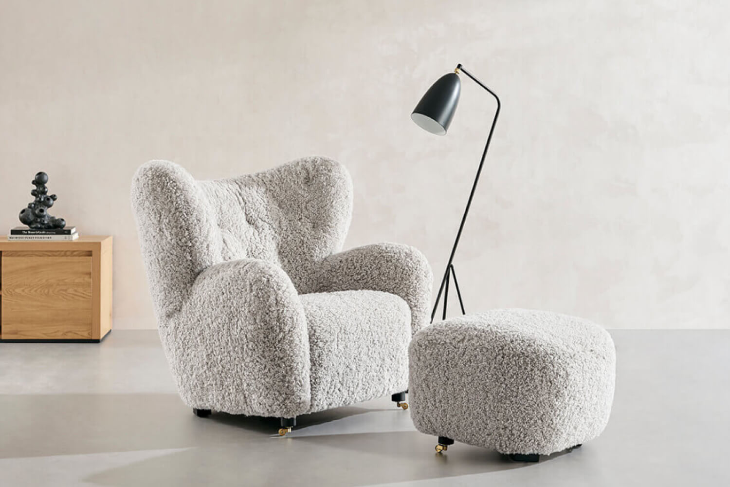 A cozy and inviting Flemming Lassen The Tired Man Lounge Chair and matching Footstool, both upholstered in genuine sheepskin, offer a luxurious seating experience. Positioned next to a sleek floor lamp and a wooden side table with a decorative sculpture.