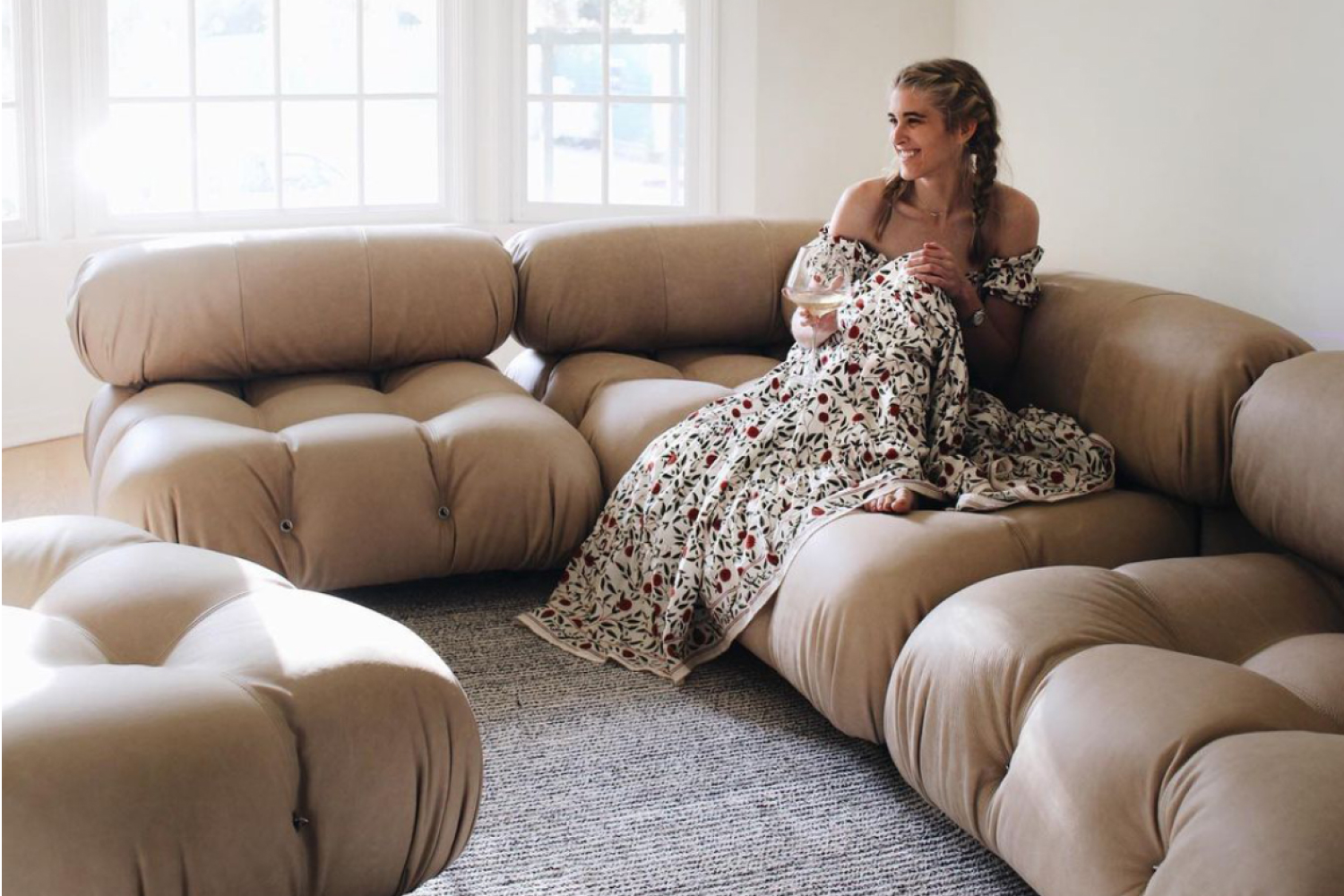 A smiling woman in a floral dress is seated on Mario Bellini modular sofa, holding a glass of wine. The room is bright with natural light, highlighting the sofa's unique design and the cozy textured grey rug beneath.
