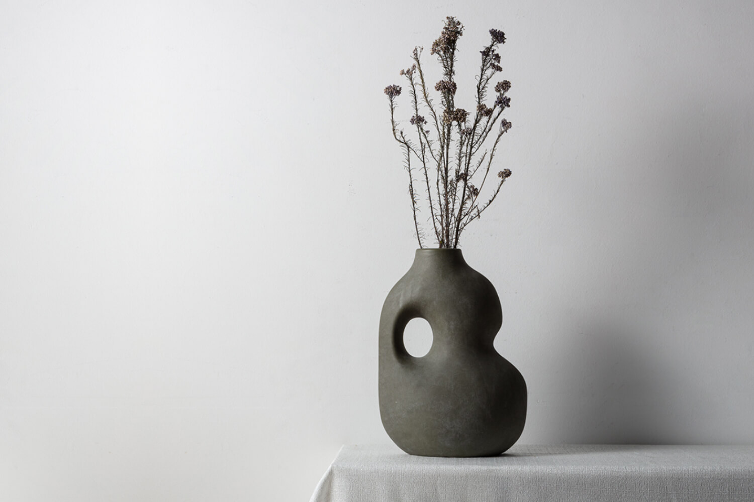 A Willow Decorative Sculptural Faded Earth Grey Rustic Wabi Sabi Ceramic Vase on a table, with slender dried flowers adding an elegant touch to its unique design.