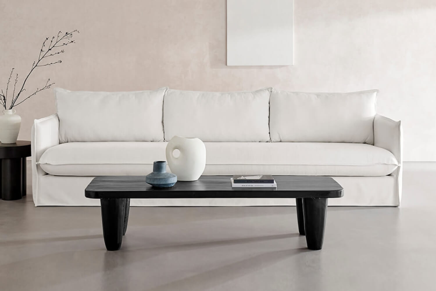 A minimalist living room with a sleek, white sofa adorned with three matching cushions. In the foreground, an EM Wabisabi Rustic Rectangular Black Reclaimed Wood Coffee Table holds a simple white vase and a book. 