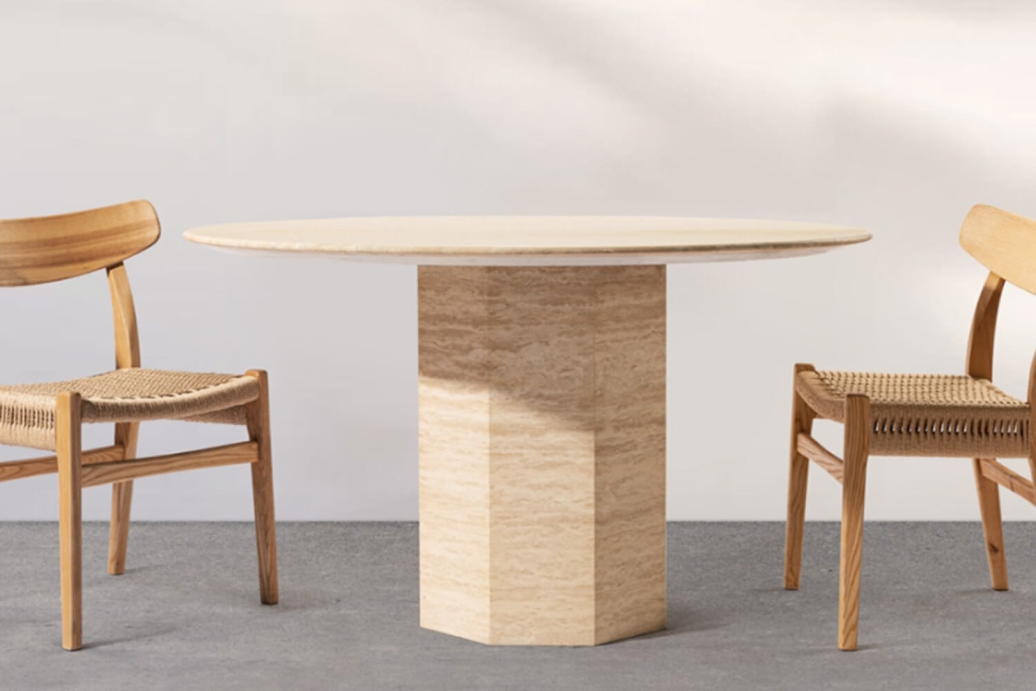 The 'Picco Round Beige Travertine Dining Table with Hexagon Base' is positioned between two woven chairs. The table's unique hexagonal base and smooth round top exhibit a natural stone pattern, offering a blend of geometric design and organic textures.